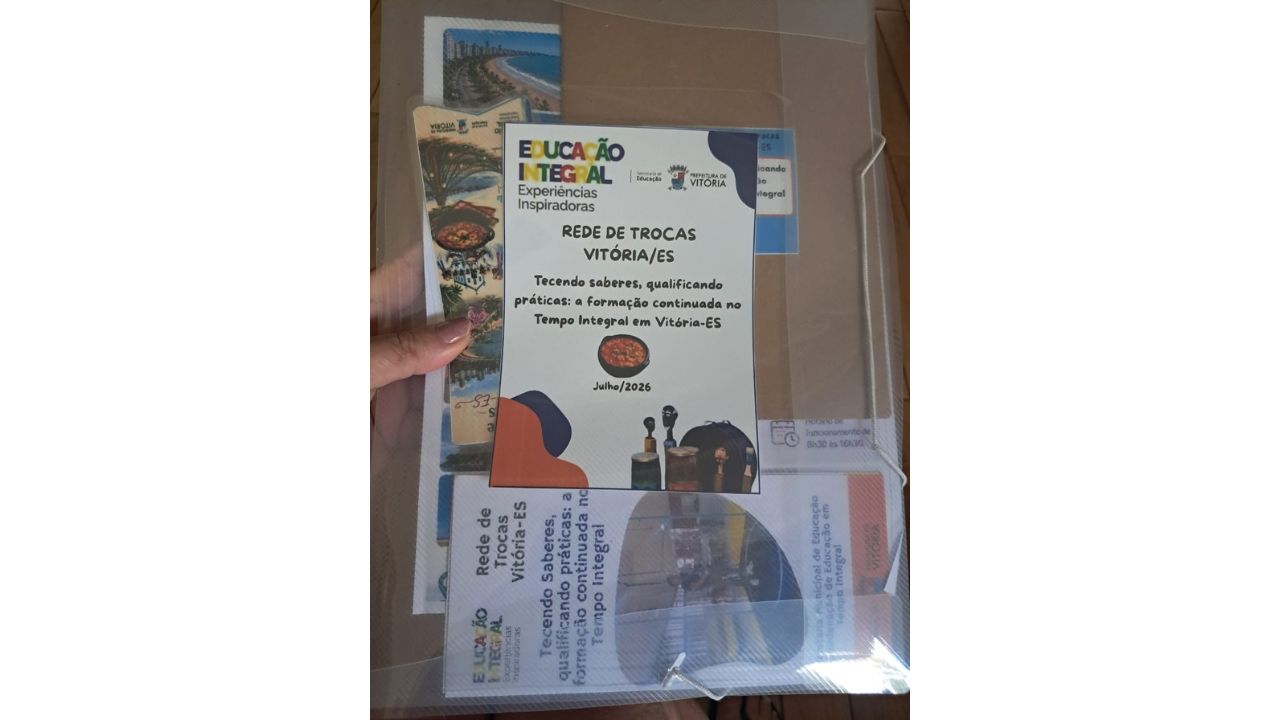 Uma mão segura uma pasta plástica transparente com documentos impressos. No centro, destaca-se um panfleto com os textos "Educação Integral", "Rede de Trocas Vitória/ES" e "Julho/2026".