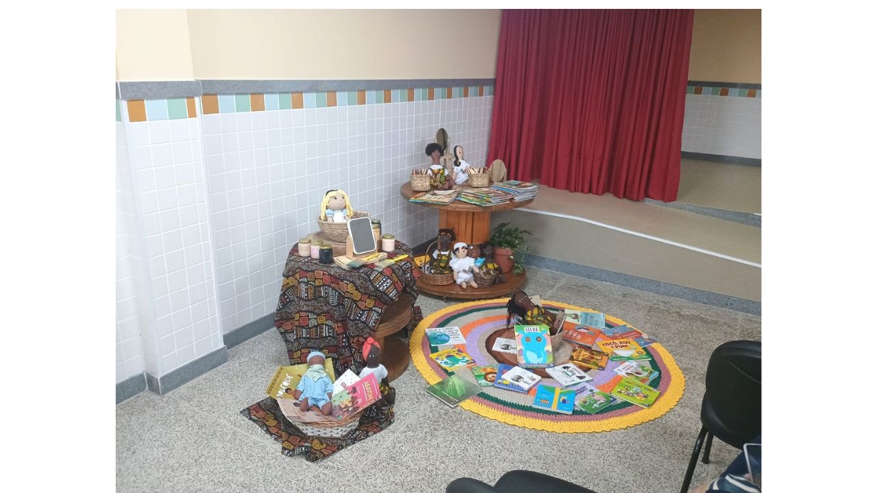 Em um ambiente educativo, carretéis de madeira e tecidos rústicos servem de suporte para bonecas de pano multirraciais, pequenos espelhos e potes. No chão, um tapete de crochê circular colorido exibe diversos livros infantis espalhados. Ao fundo, nota-se uma parede com azulejos brancos e uma faixa colorida, além de uma cortina vermelha à direita.