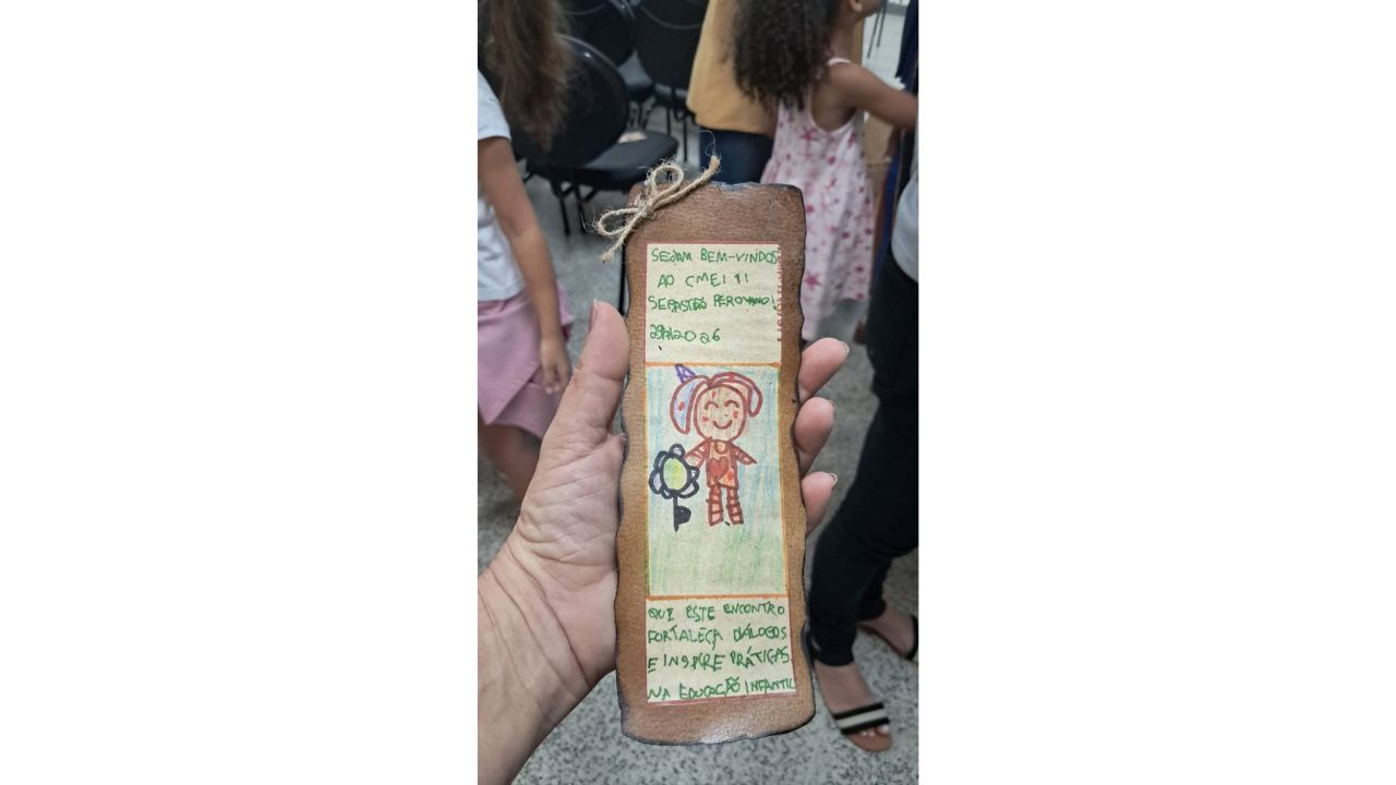 Uma mão segura um marcador de página artesanal retangular. O marcador exibe um texto manuscrito em verde - "Sejam bem-vindos ao Cmei TI Sebastião Perovano. Que esse encontro fortaleça diálogos e inspire práticas na Educação Infantil" - e o desenho de uma figura infantil com uma flor. Ao fundo, pernas de adultos e crianças em movimento são vistas em um salão com cadeiras pretas.