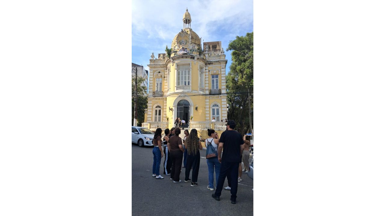 Um grupo multirracial de cerca de 10 pessoas aparece de costas em uma rua asfaltada. Elas estão em pé, de costas e de perfil, e vestem trajes casuais. O foco central é um casarão histórico amarelo, com adornos brancos, varandas, porta em arco e uma cúpula ornamentada no telhado. Ao fundo e nas laterais, há carros estacionados, árvores e postes de energia elétrica.