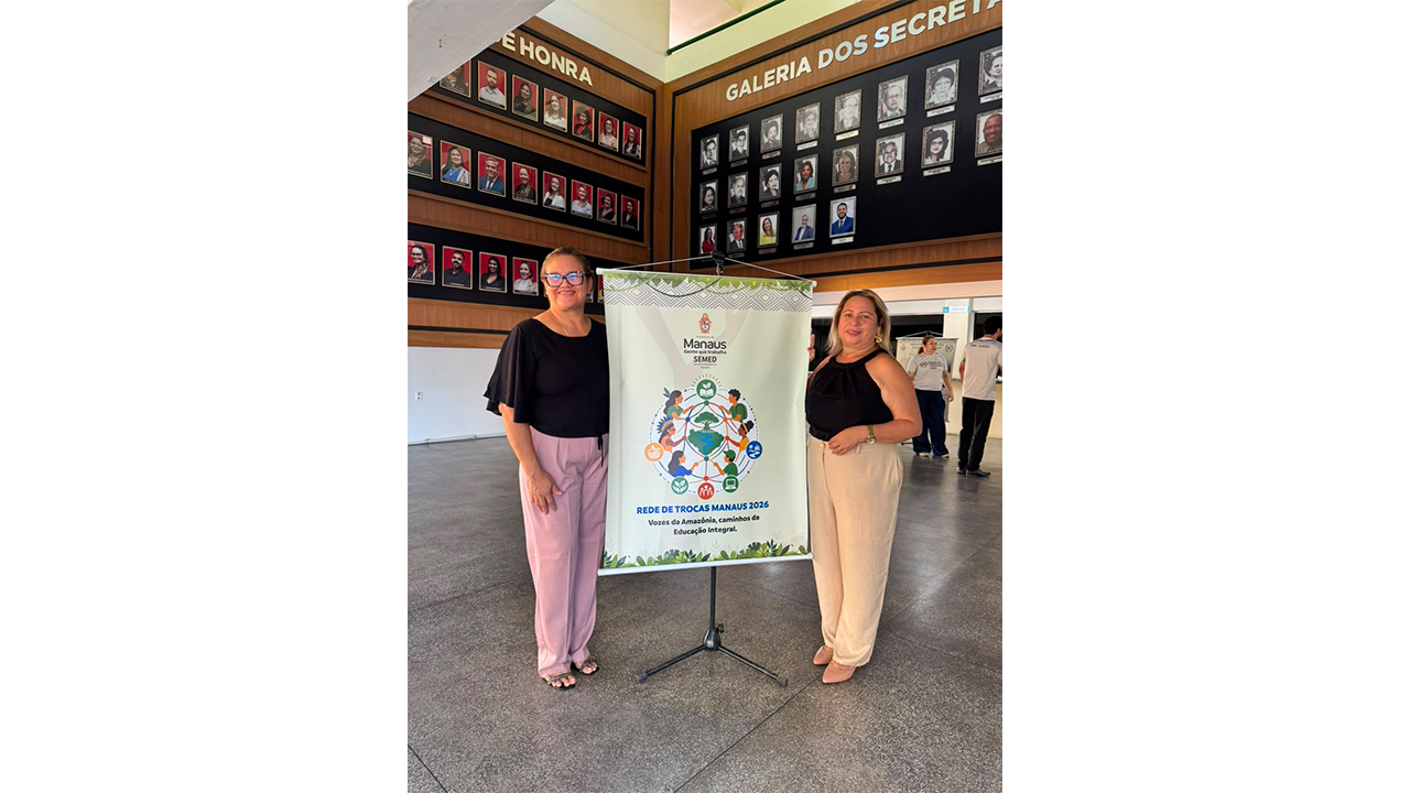 Duas mulheres estão em pé, usando roupas casuais, ao lado de um banner da "Rede de Trocas Manaus 2026". Ao fundo, destaca-se um grande painel de madeira contendo diversos retratos emoldurados de rostos, sobre um piso cinza liso.
