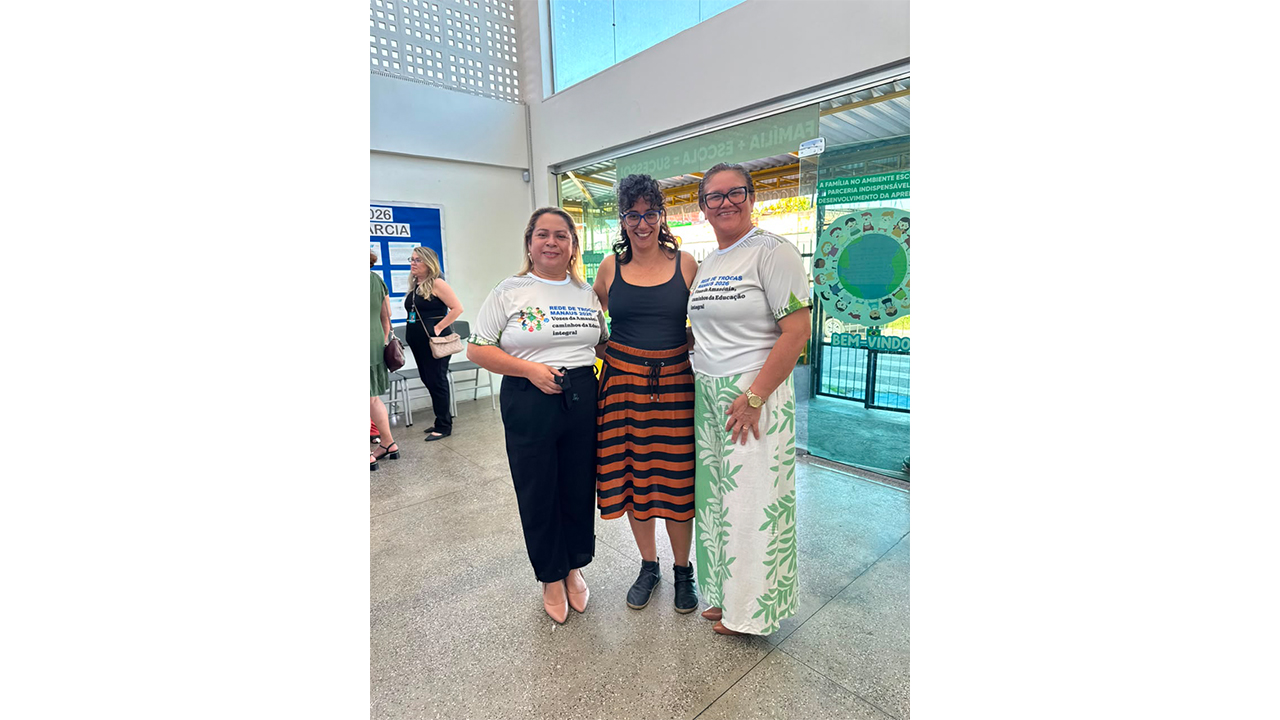Três mulheres estão em pé, lado a lado, sorrindo. Duas vestem camisetas brancas semelhantes, e a do centro usa blusa preta e saia listrada. O fundo revela portas de vidro transparente, permitindo ver a estrutura externa, a rua e algumas pessoas passando.