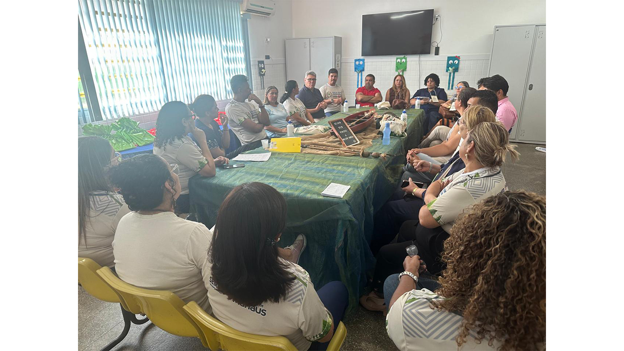 Um grupo multirracial de cerca de vinte pessoas está sentado em roda ao redor de uma grande mesa. O centro da mesa é forrado com tecido verde e exibe uma miniatura de canoa de madeira. O cenário possui cadeiras amarelas em primeiro plano, paredes brancas com azulejos claros e uma TV fixada ao fundo.