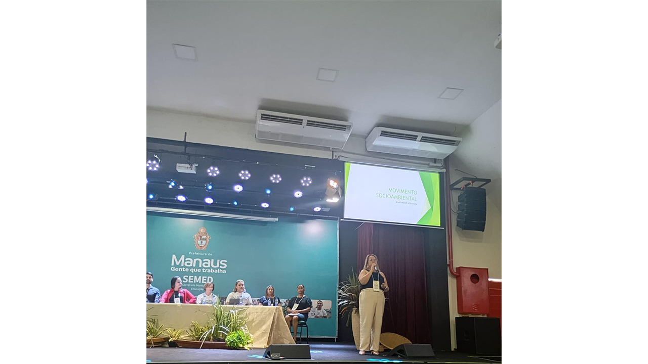 Uma mulher em pé, à direita, fala ao microfone, enquanto seis pessoas estão sentadas à esquerda, atrás de uma mesa. Destacam-se um painel verde com o texto "Prefeitura de Manaus SEMED" e um telão superior projetando uma apresentação. O fundo inclui teto branco, aparelhos de ar-condicionado e extintores de incêndio.