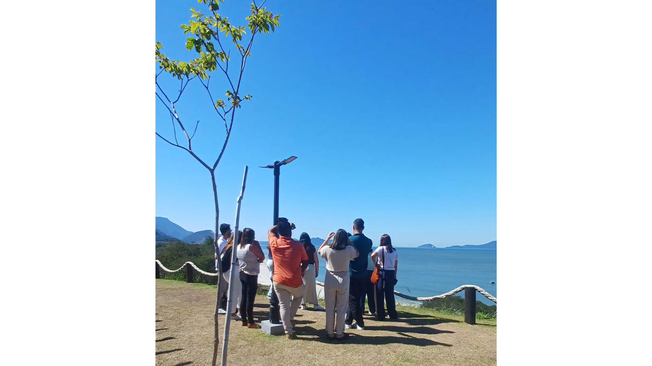 Em um dia de céu azul, um grupo de aproximadamente oito adultos está de costas, observando o mar e pequenas ilhas ao longe. O espaço conta com um gramado e um guarda corpo de cordas brancas, apoiadas em pequenos tocos de madeira escura.