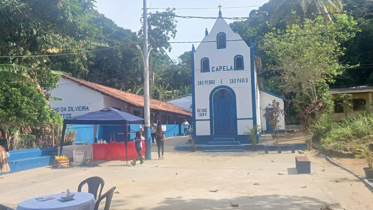 Em uma praça arborizada, uma pequena capela branca e azul está ao centro, com as inscrições "São Pedro e São Paulo, desde 1884". Algumas pessoas estão sentadas ou em pé no entorno, próximas a mesas e barracas. Ao redor, há árvores e construções simples, além de postes e fios de energia.