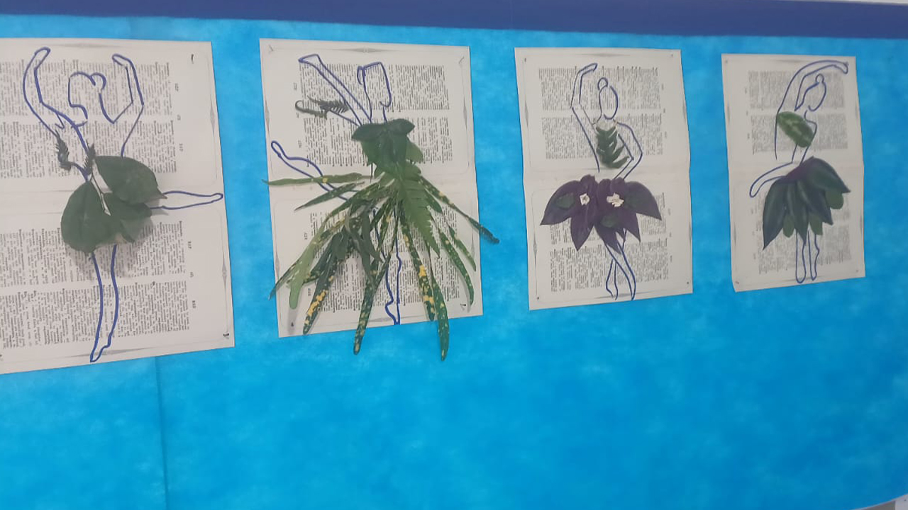 Painel escolar azul com quatro trabalhos artísticos alinhados. Cada composição apresenta o contorno de uma figura humana desenhado de caneta sobre páginas impressas, com folhas verdes e flores naturais em tonalidades escuras formando partes das roupas, como saias e vestidos.