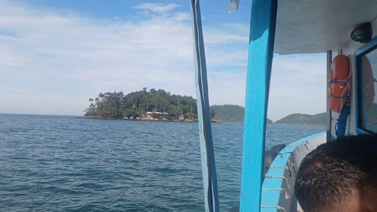 Vista a partir de uma embarcação em movimento sobre o mar. Em primeiro plano, aparecem parte da estrutura branca com detalhes em azul do barco e a cabeça de uma pessoa; ao centro e ao longe, vê-se uma pequena ilha coberta por vegetação.