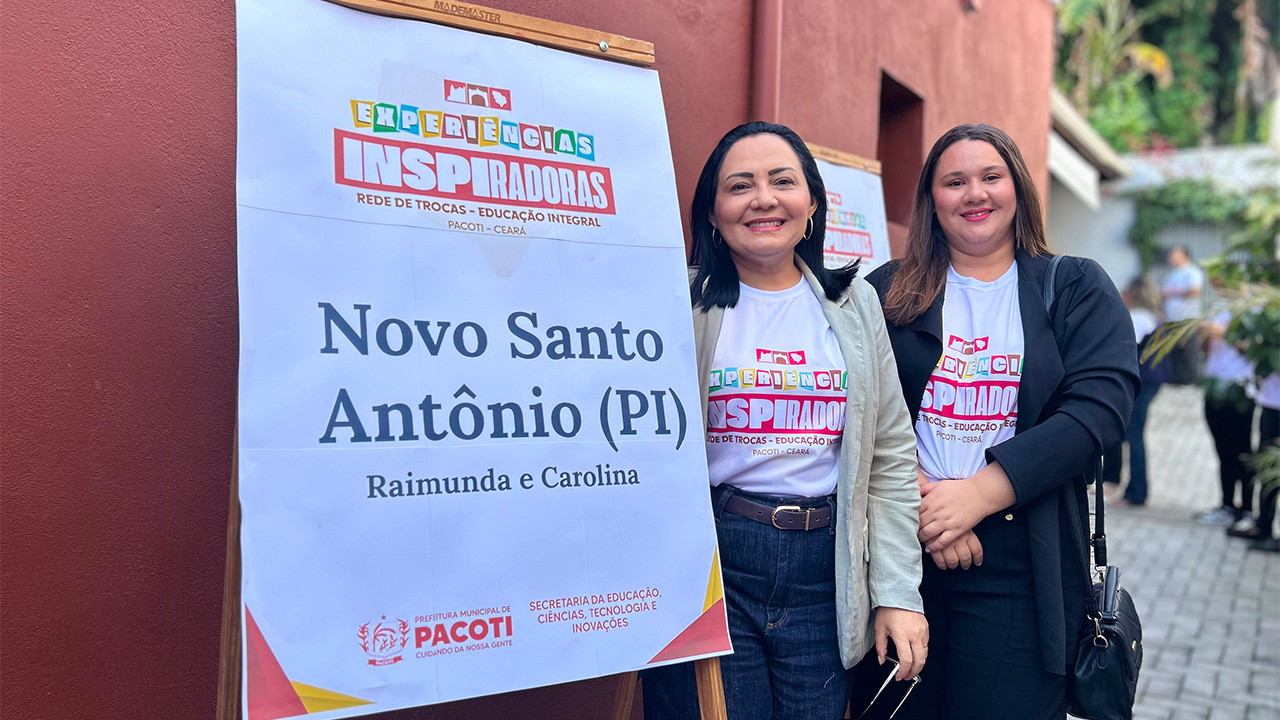 Duas mulheres usam calças jeans escuras e camisetas brancas iguais com o nome "Experiências inspiradoras" posam lado a lado diante de um cavalete de madeira com os textos “Experiências Inspiradoras – Rede de Trocas – Educação Integral” e “Novo Santo Antônio (PI) – Raimunda e Carolina”. Ao fundo, há outras pessoas, vegetação e uma grande parede vermelha