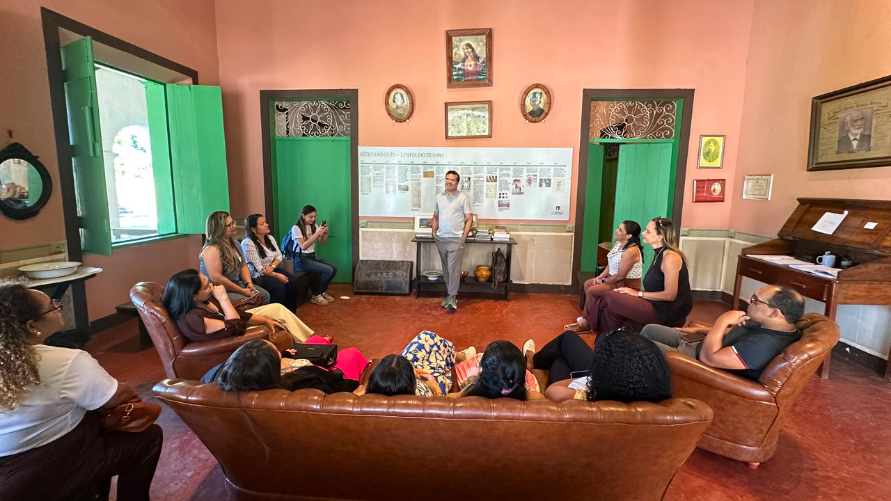 Ambiente interno com paredes rosadas, duas portas e uma janela verdes. Um homem fala em pé para um grupo multirracial de 12 adultos sentados em sofás e cadeiras de couro marrom ao redor. Ao fundo, há quadros, móveis antigos e um painel com uma linha do tempo