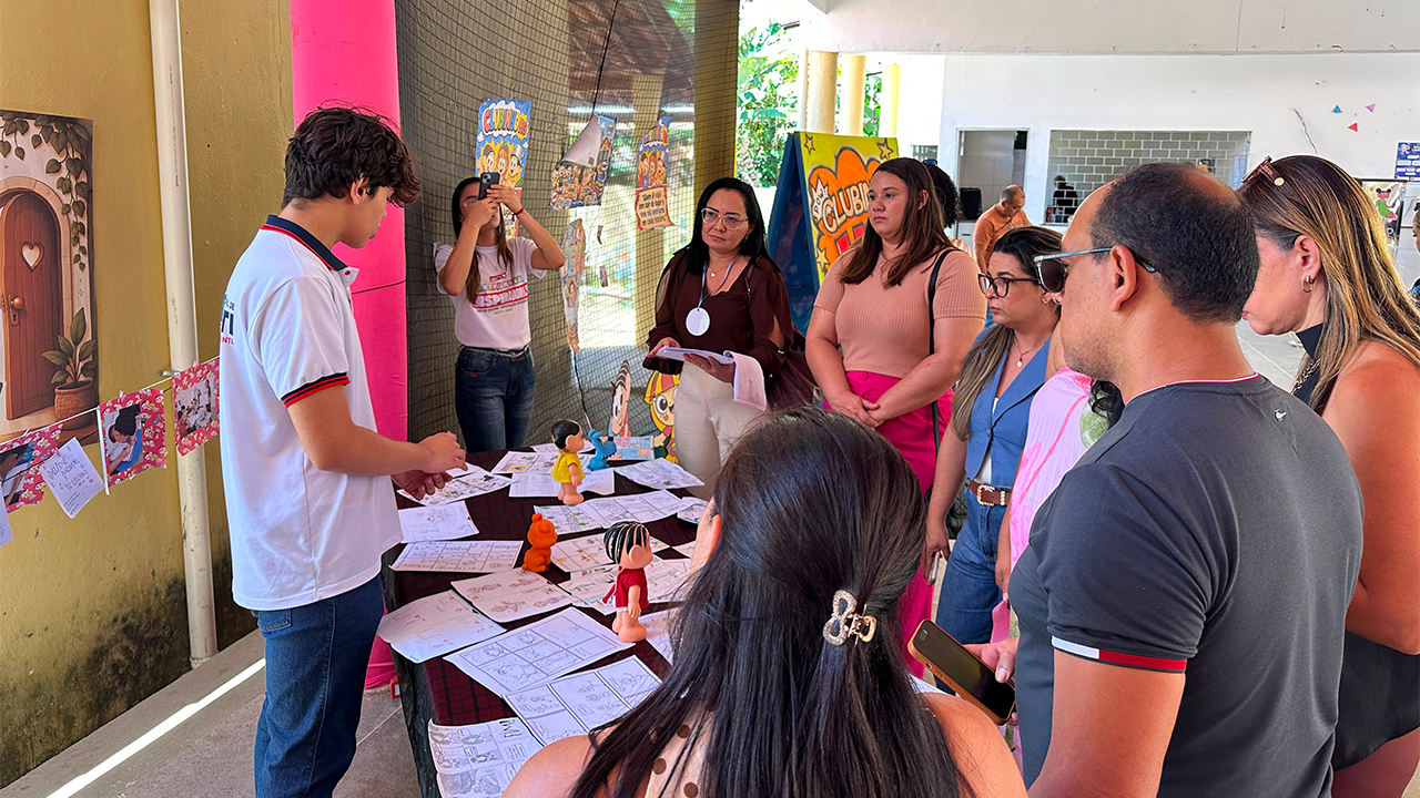 Espaço escolar de exposição, com uma mesa coberta por desenhos, histórias em quadrinhos e bonecos da Turma da Mônica, como Mônica e Magali. Um estudante, vestindo camiseta branca e de costas para a câmera, apresenta os trabalhos a um grupo de adultos reunidos ao redor. Ao fundo, há painéis coloridos e elementos decorativos.