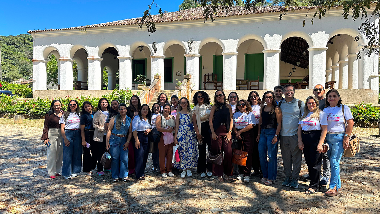 Um grupo multirracial de cerca de vinte e cinco adultos posa lado a lado, usando roupas casuais e algumas camisetas padronizadas com o título "Experiências inspiradoras". Ao fundo, há um grande casarão branco, com uma sequência de arcos, portas verdes e luminárias pendentes. Ao redor, há jardim, árvores e piso de pedras.