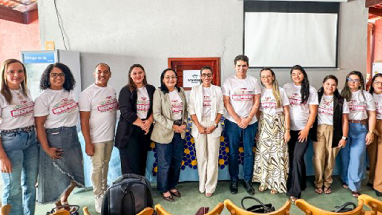 Grupo multirracial de doze adultos posa lado a lado. Todas as pessoas vestem camisetas brancas com os dizeres "Experiências inspiradoras", complementadas por calças jenas ou sociais e blazers. Ao fundo, há uma tela de projeção desligada e fixada na parede branca.
