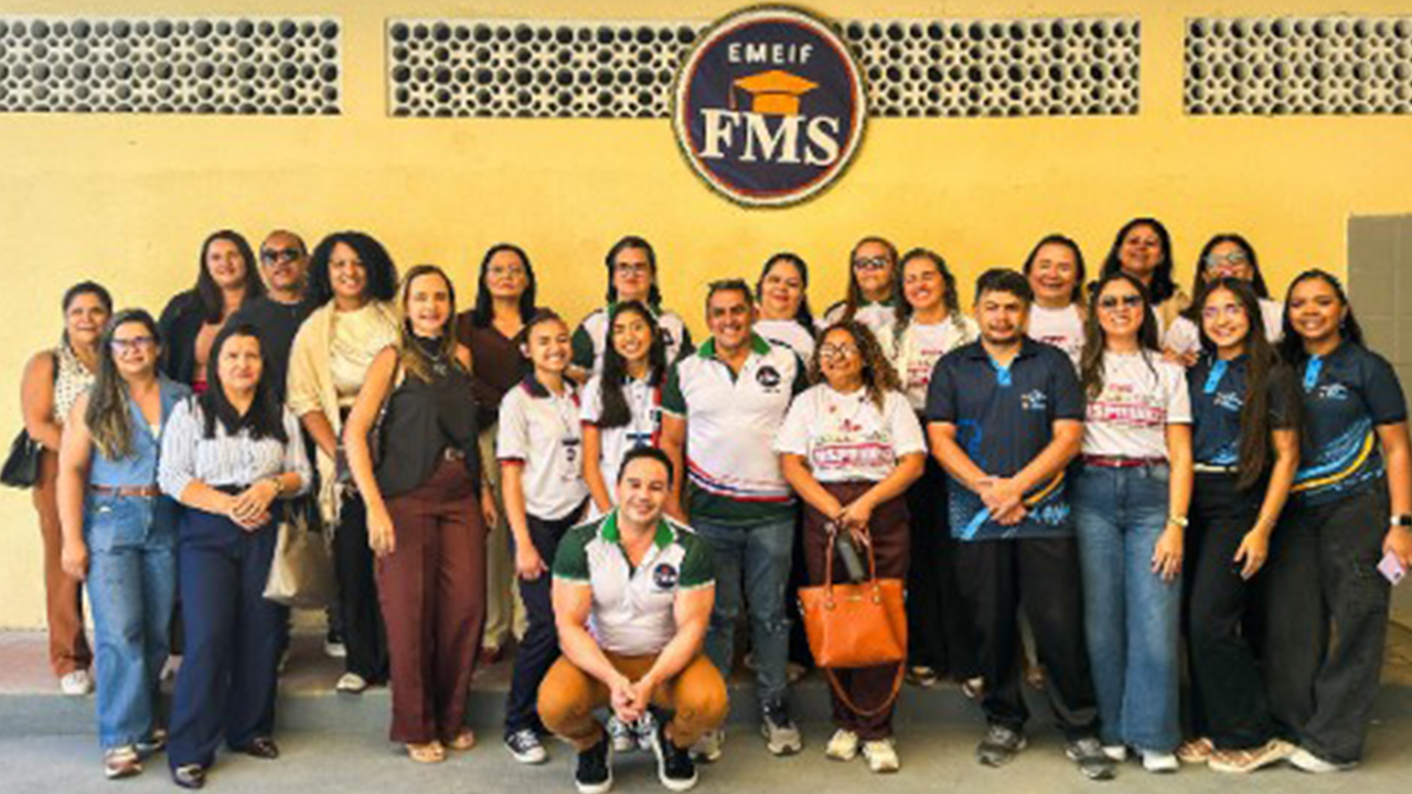 Um grupo multirracial de cerca de 25 pessoas, entre adultos e jovens, em trajes casuais, posa lado a lado. Atrás deles, em uma parede amarela com blocos vazados, uma placa azul circular exibe o texto “EMEIF FMS” e o desenho de um capelo laranja.