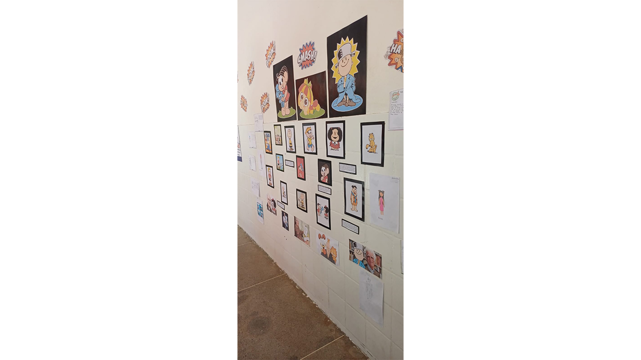 Parede escolar de azulejos brancos decorada com trabalhos e imagens de personagens de histórias em quadrinhos. Há desenhos, cartazes e figuras emolduradas distribuídos em diferentes alturas, além de ilustrações de onomatopeias, como "smash" e "pow". Também é possível identificar um desenho do Menino Maluquinho e da Mônica.