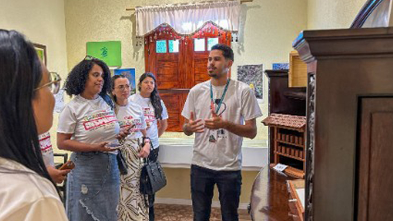 Grupo multirracial composto por quatro pessoas acompanha a fala de um homem em pé, que gesticula durante a explicação. As três mulheres da foto usam camisetas brancas iguais, com os dizeres "Experiências inspiradoras". As paredes são de um amarelo claro com um papel de parede de arabescos verdes próximo ao teto. Ao redor, há móveis de madeira, cortinas e objetos decorativos.
