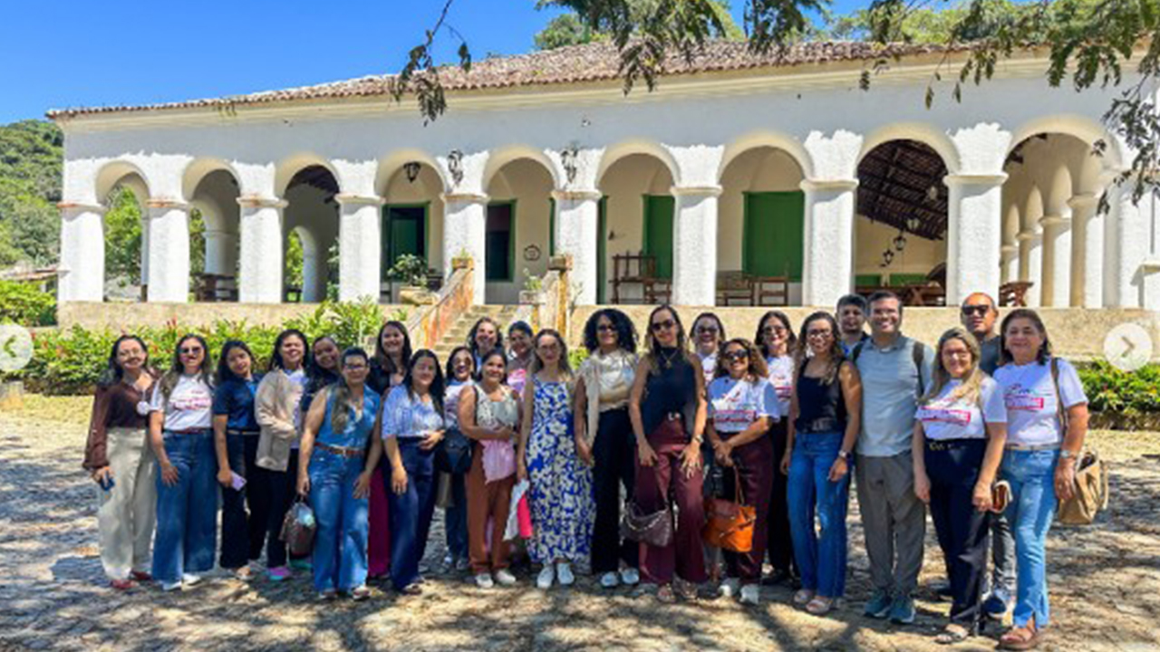 Um grupo multirracial de cerca de vinte e cinco adultos posa lado a lado, usando roupas casuais e algumas camisetas padronizadas com o título "Experiências inspiradoras". Ao fundo, há um grande casarão branco, com uma sequência de arcos, portas verdes e luminárias pendentes. Ao redor, há jardim, árvores e piso de pedras.