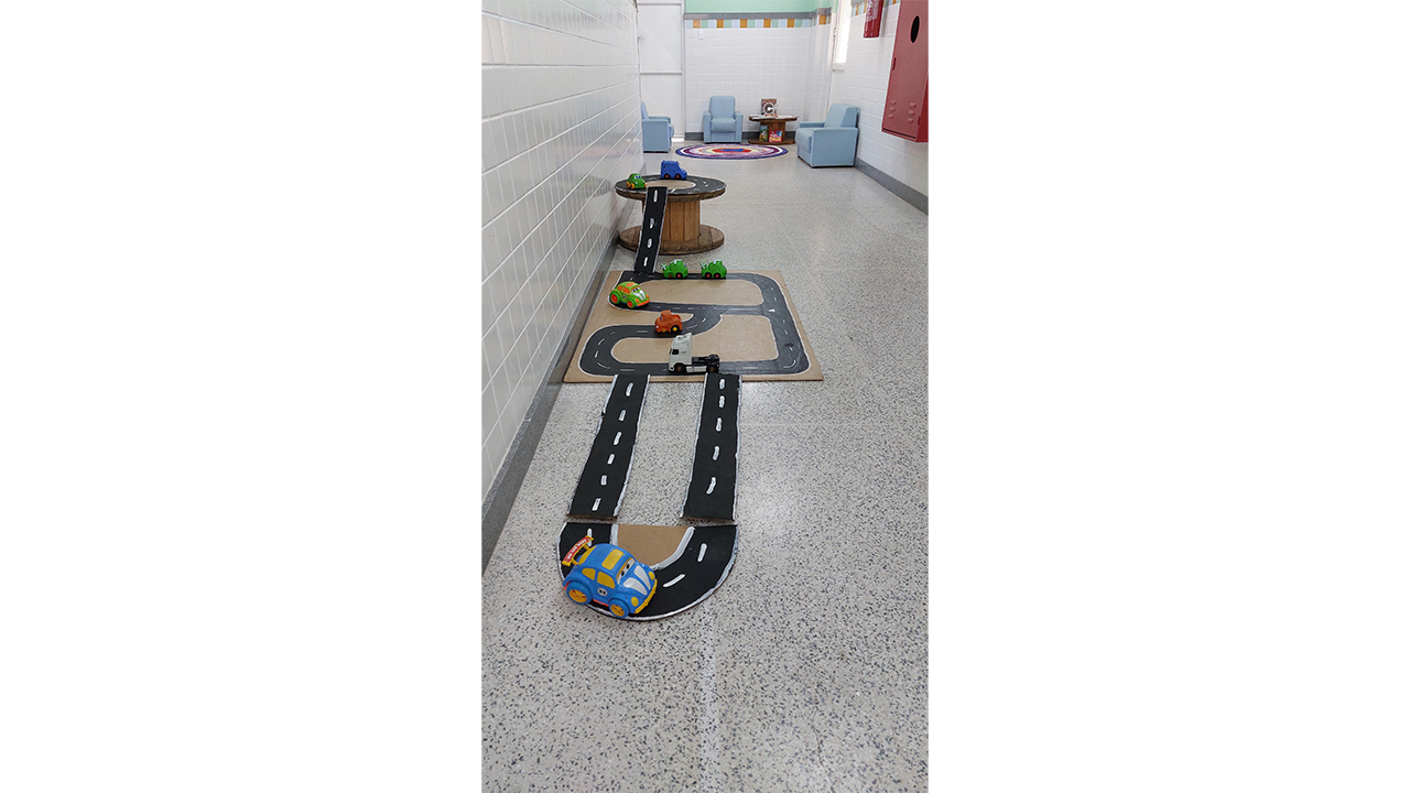 No chão de um corredor escolar de paredes brancas de azulejos e piso cinza, há uma pista de carrinhos feita de papelão preto, com faixas brancas pintadas e carrinhos de plástico espalhados. Ao fundo, vê-se uma área de leitura com pequenas poltronas azuis. À direita, há um extintor de incêndio vermelho.
