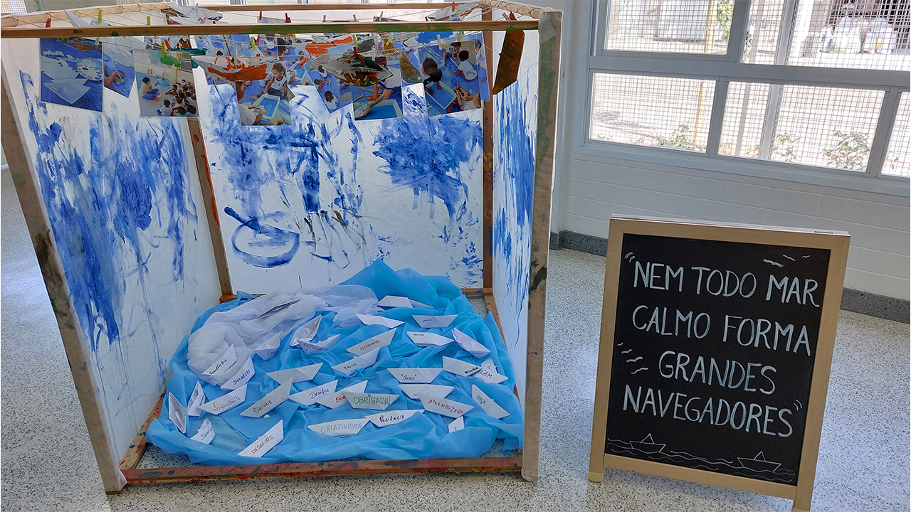 Uma estrutura de madeira em formato de cubo exibe barcos de papel branco sobre um tecido azul e fotos penduradas em um varal. Ao lado, uma lousa de cavalete com fundo preto exibe o texto: "Nem todo mar calmo forma grandes navegadores”. Ao fundo, há janelas com grades.