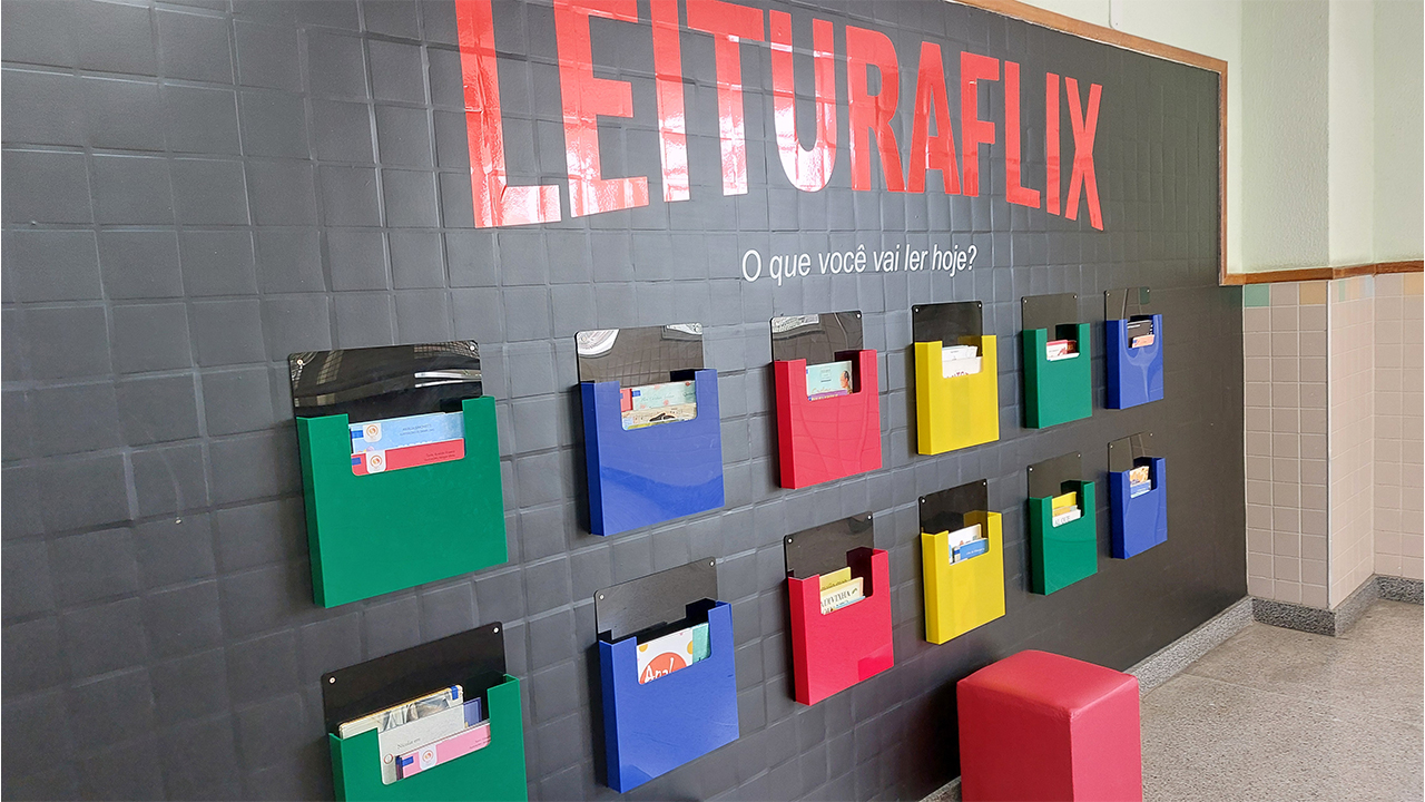Mural escolar em uma parede preta. No topo, letras vermelhas e brancas formam o texto "LEITURAFLIX - O que você vai ler hoje?". Abaixo, dez nichos de acrílico nas cores verde, azul, vermelha e amarela exibem diversos livros infantis.