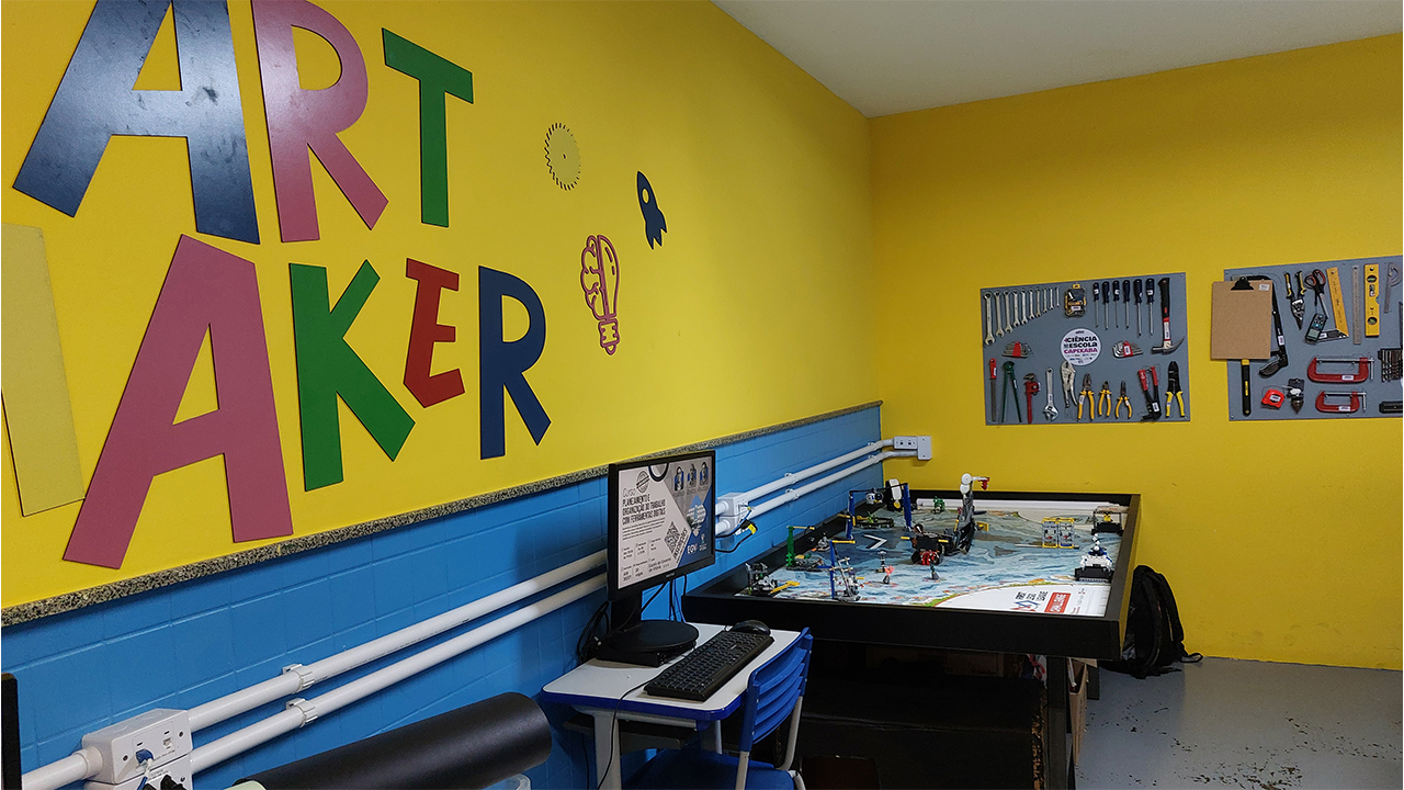 Em um laboratório escolar, a parede amarela exibe um grande letreiro colorido "ART MAKER". O espaço contém uma mesa preta com blocos de montar, uma mesa com um computador ao lado e dois painéis cinzas de parede que organizam ferramentas. A parte inferior da parede é azul e o piso é cinza.