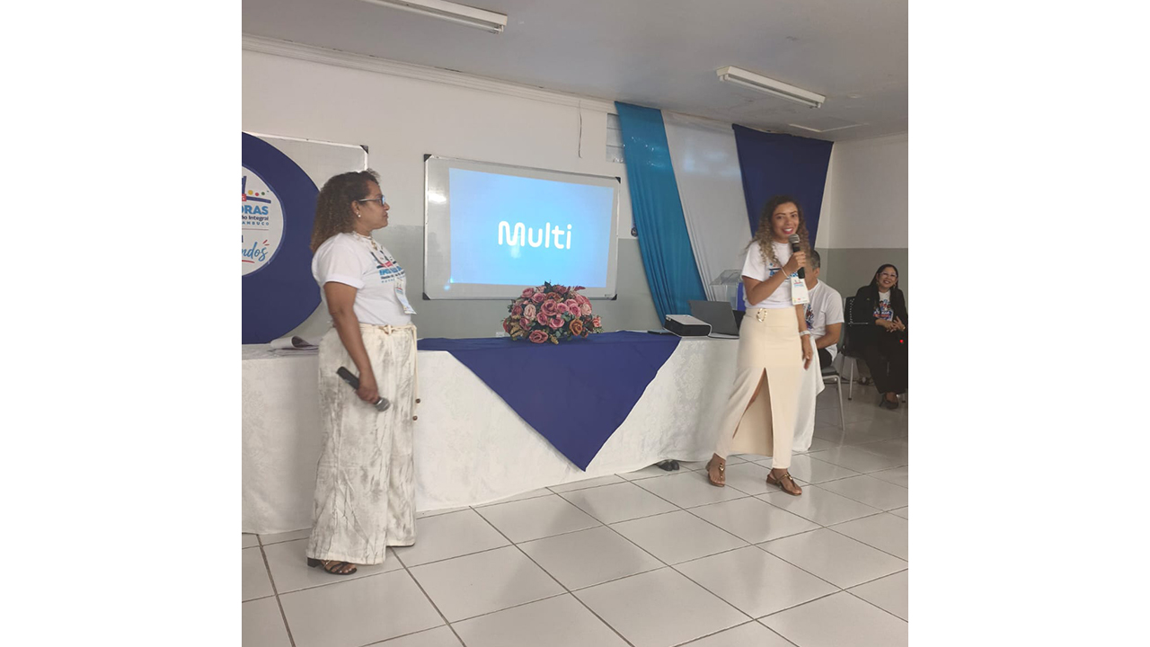 Duas mulheres estão em pé à frente de uma mesa em uma sala. A da direita, de saia bege e camiseta branca, fala ao microfone, enquanto a da esquerda, de calça clara estampada, a observa, segurando outro microfone. Na parede atrás delas, um projetor exibe a palavra "Multi", em um fundo azul. O ambiente possui piso de cerâmica branca, uma mesa com toalha azul e branca, contendo um arranjo floral, e tecidos decorativos pendurados ao fundo.