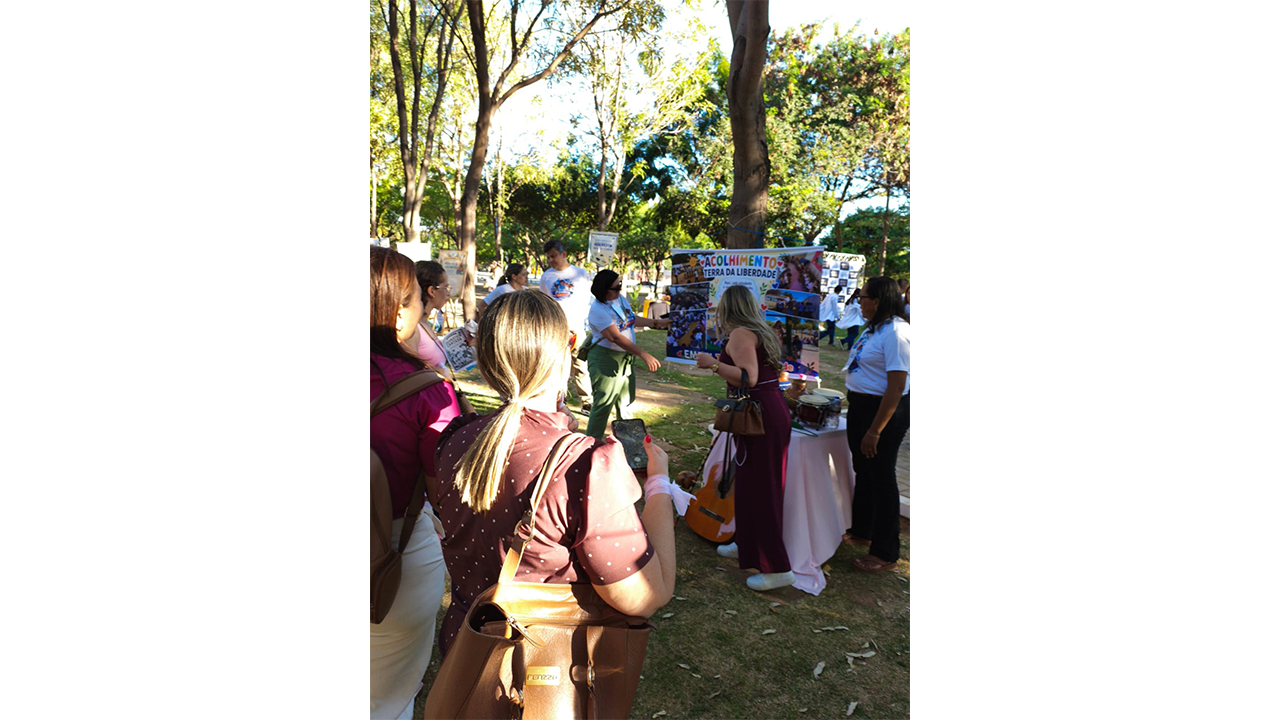 Em um ambiente externo, diversas pessoas caminham e conversam. Em primeiro plano, uma mulher vista de costas, de blusa marrom. No centro, o destaque é um banner repleto de fotos e o texto "Acolhimento: Terra da liberdade", pendurado em uma árvore, ao lado de uma mesa forrada de rosa, com pequenos tambores e um violão escorado. O fundo exibe um gramado com folhas secas e troncos de árvores altas.