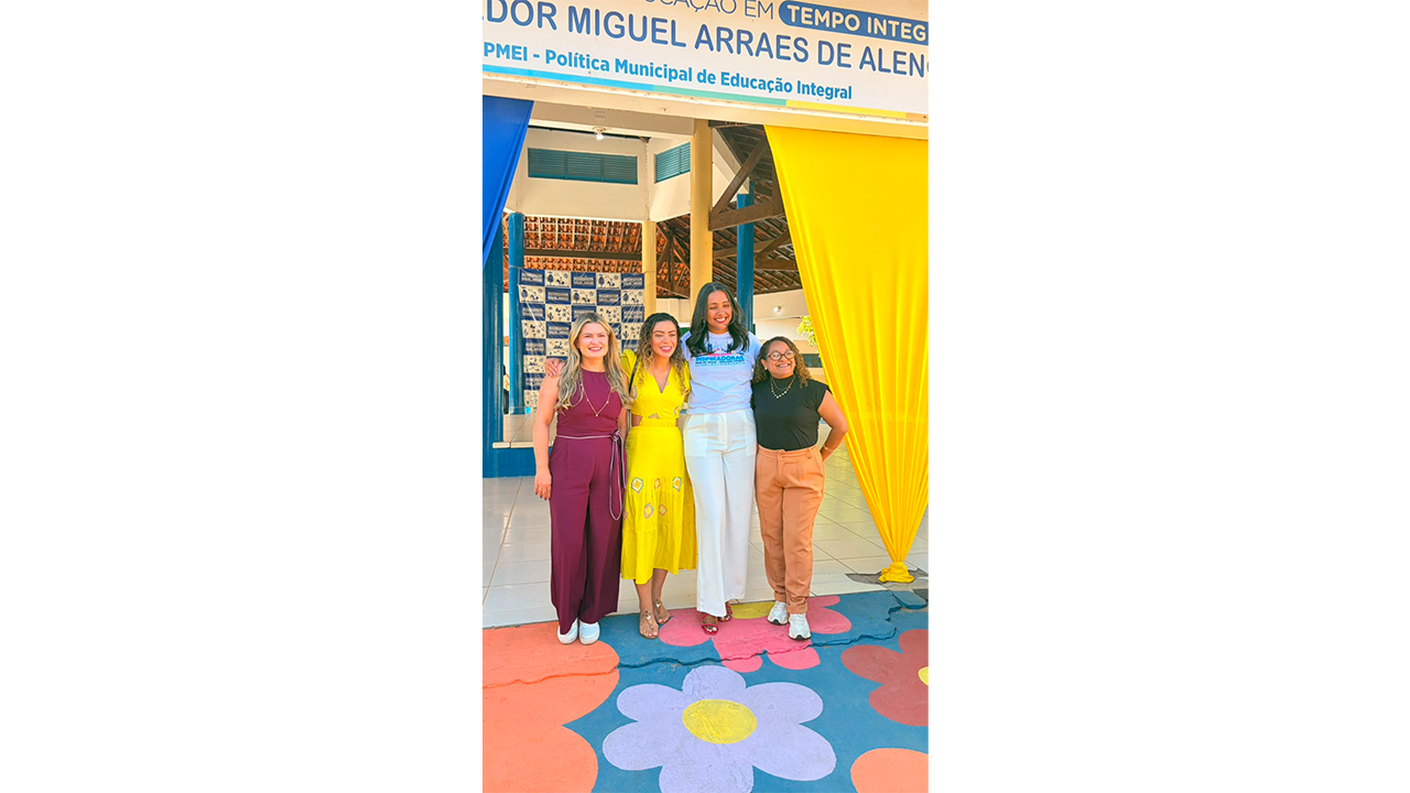 Um grupo multirracial de quatro mulheres posa junto na entrada de uma escola. Elas estão em pé, lado a lado, vestindo trajes casuais em tons de bordô, amarelo, branco e preto. Acima delas, uma placa exibe textos parcialmente visíveis e a frase "PMEI - Política Municipal de Educação Integral". O piso é decorado com grandes flores pintadas, e o fundo mostra a estrutura coberta de um prédio, ladeada por tecidos azuis e amarelos.