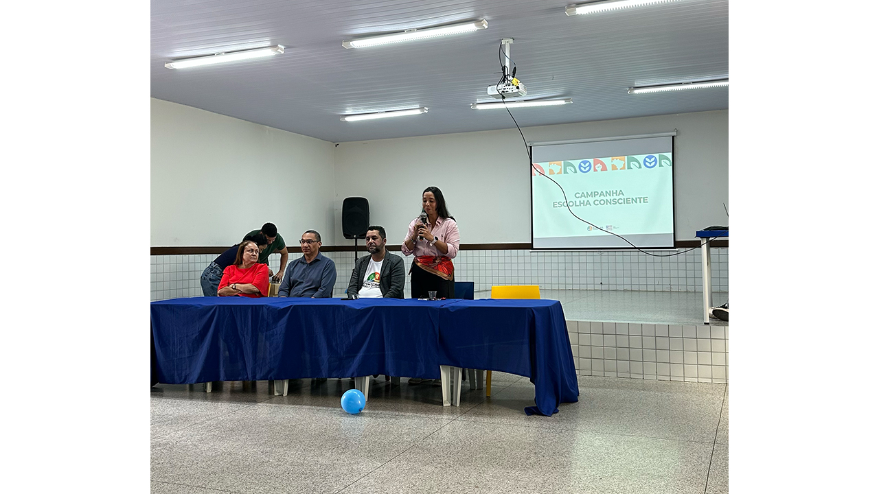 Em uma sala de paredes brancas e piso claro, três adultos estão sentados em uma mesa com toalha azul à frente da sala, enquanto uma mulher, em pé, fala ao microfone. Atrás do grupo, uma tela de projeção tem o título “Campanha Escolha Consciente”.