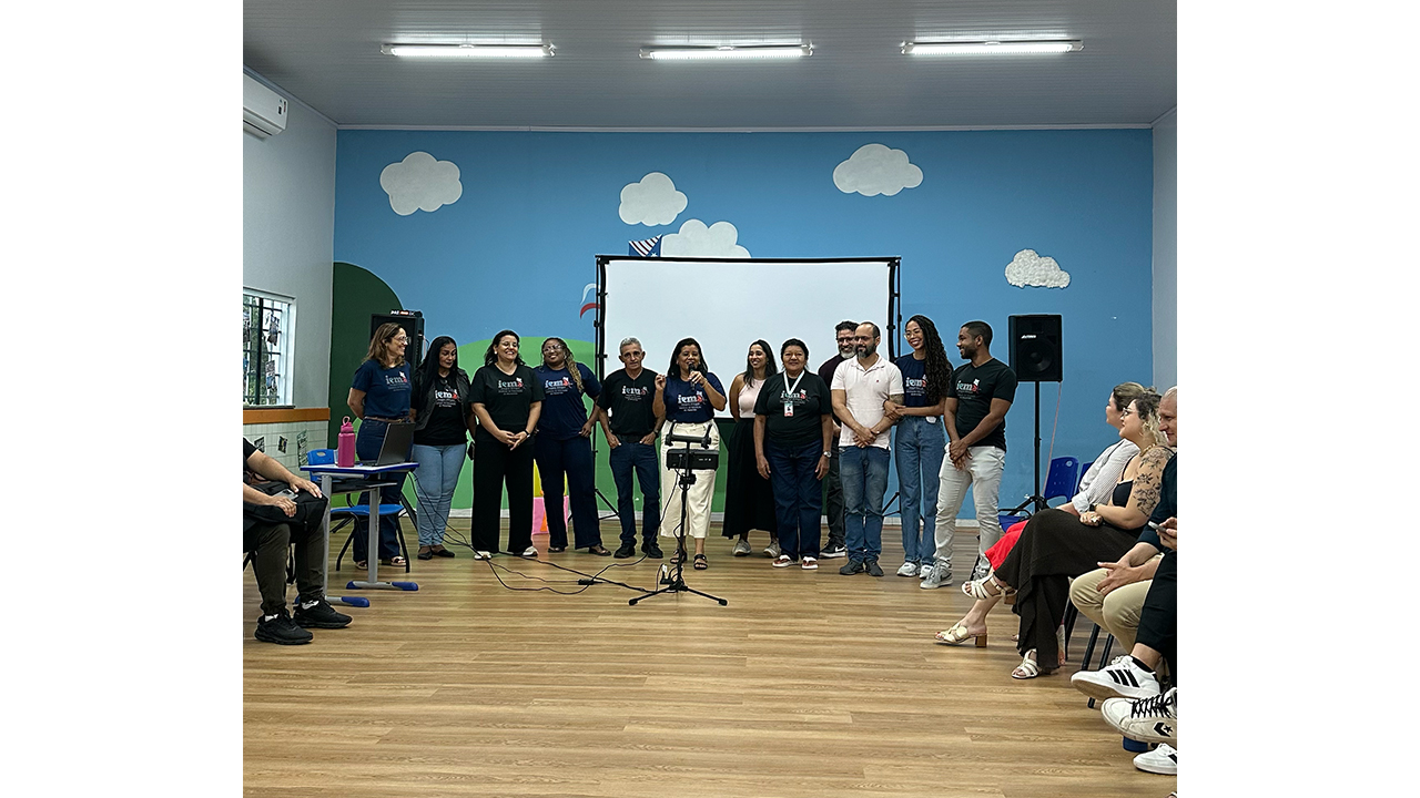Sala ampla com adultos reunidos em semicírculo e um grupo multirracial de 12 pessoas em pé diante de uma tela de projeção. Uma mulher, ao centro, fala ao microfone, enquanto os demais acompanham a apresentação. Ao fundo, há uma parede azul com nuvens decorativas e caixas de som. O piso é de madeira clara.