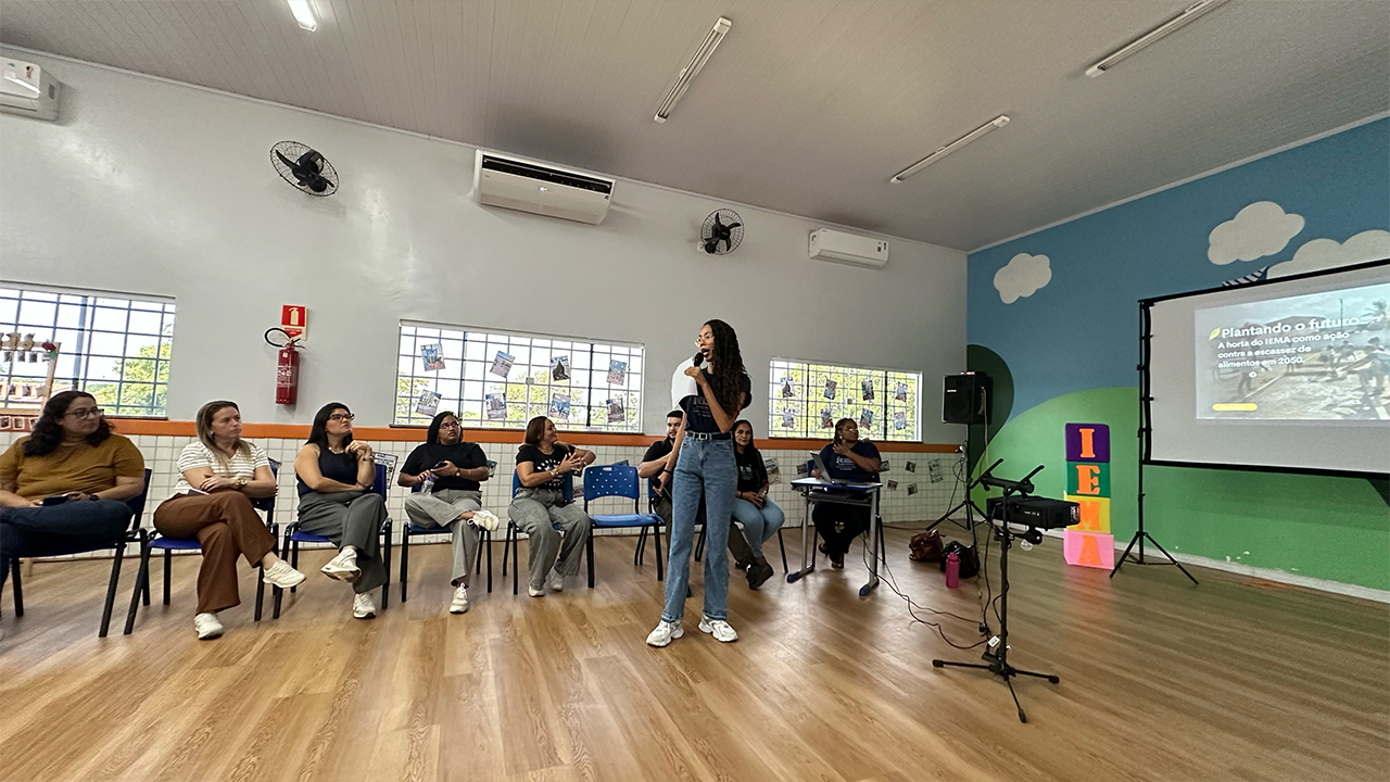Grupo multirracial de oito adultos disostos em semicírculo e sentados em cadeiras de plástico azuis acompanha apresentação em um sala de reunião. Uma jovem, em pé, fala ao microfone diante de uma projeção com o título “Plantando o futuro”. Ao fundo, há janelas com fotografias coladas no vidro, aparelhos de ar-condicionado, ventiladores pretos e uma parede com fundo azul, nuvens brancas e montanhas verdes, além de uma torre de cubos coloridos com as letras I, E, M e A em laranja.