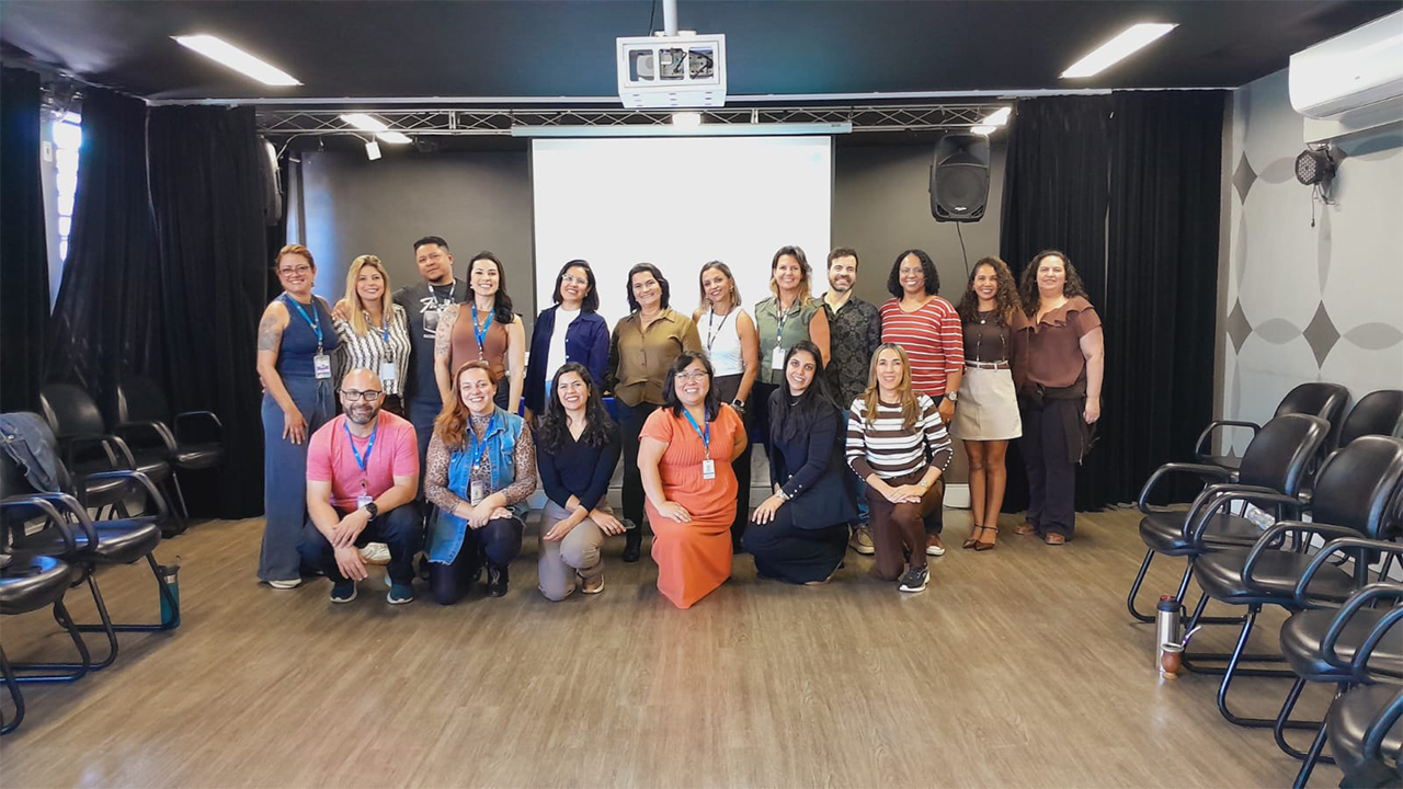 Em uma sala de apresentação, um grupo multirracial formado por cerca de dezoito adultos posa para a fotografia, alguns em pé e outros agachados. As pessoas usam roupas casuais. Ao fundo, há tela de projeção branca, cortinas pretas, caixas de som e fileiras de cadeiras pretas. Há cortinas pretas nas janelas do lado esquerdo. O piso da sala é de madeira clara.