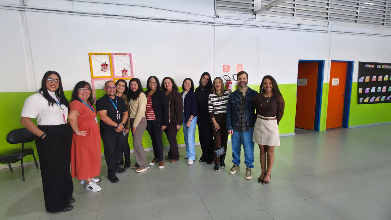 Em um corredor amplo, um grupo multirracial de 12 adultos posa lado a lado, sorrindo. O espaço conta com paredes brancas com a metade inferior na cor verde limão, o piso é cinza, as portas são laranjas com batentes azuis, e há um mural preto com trabalhos pedagógicos coloridos pendurado na parede à direita.