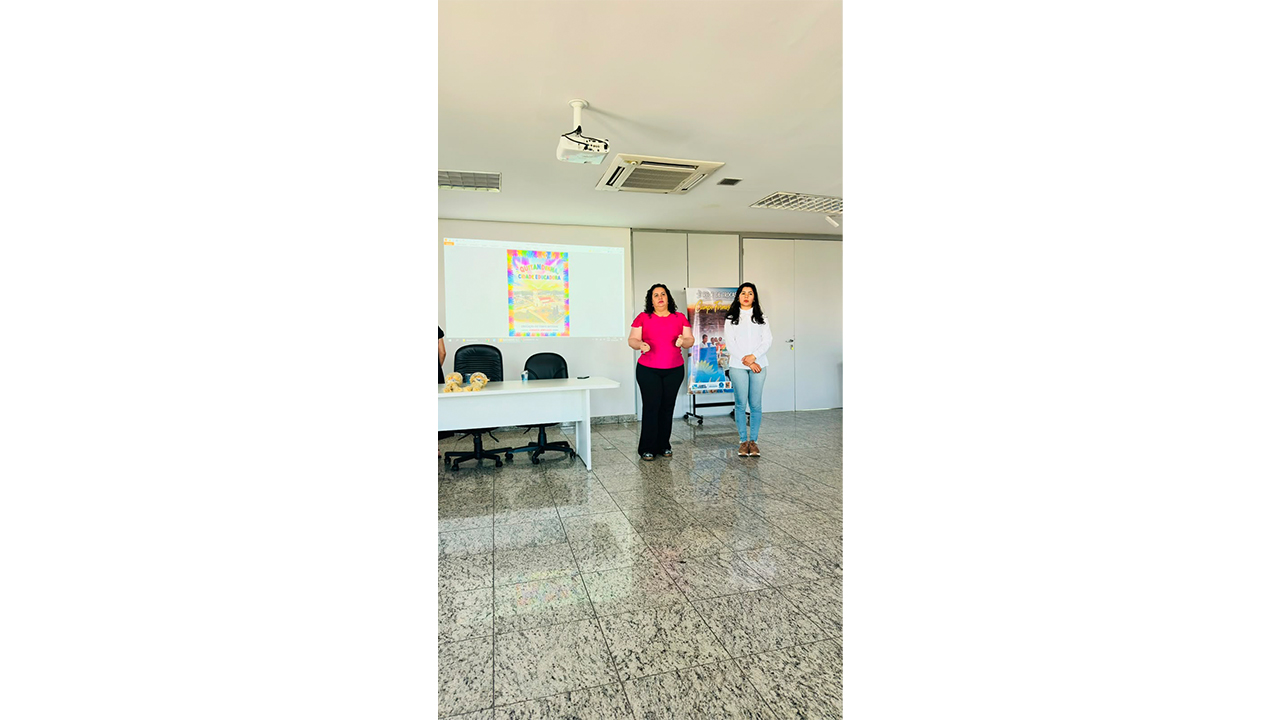Sala de apresentação com duas mulheres em pé diante de uma projeção. Uma veste blusa rosa e calça preta; a outra, blusa branca e calça jeans. Na tela aparece “Quitandinha Cidade Educadora”, e ao lado há um painel da “Rede de Trocas”. O ambiente é bem iluminado, com uma mesa branca com duas cadeiras pretas, um piso de granito cinza e projetor branco no teto.