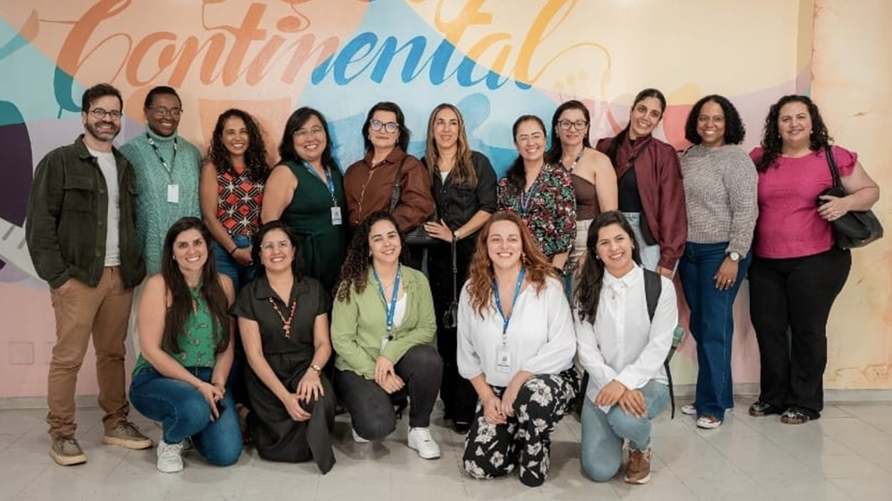 Grupo multirracial de 16 adultos posa para foto. Enquanto alguns estão em pé, outros agancham-se à frente. Todos usam roupas casuais e sociais de diversas cores. O chão é claro, enquanto a parede ao fundo tem várias cores: laranja, azul, amarelo, verde e roxo. É possível ler a palavra "continental" escrita de forma estilizada na arte da parede.