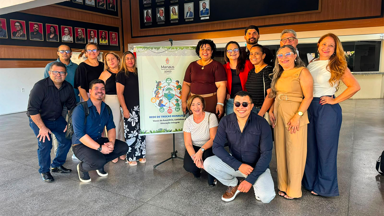 Um grupo multirracial de quinze pessoas, algumas em pé e outras agachadas, posa com roupas casuais ao lado de um tripé com banner. Ao fundo, há um piso liso e um painel de madeira escura contendo diversos retratos emoldurados.