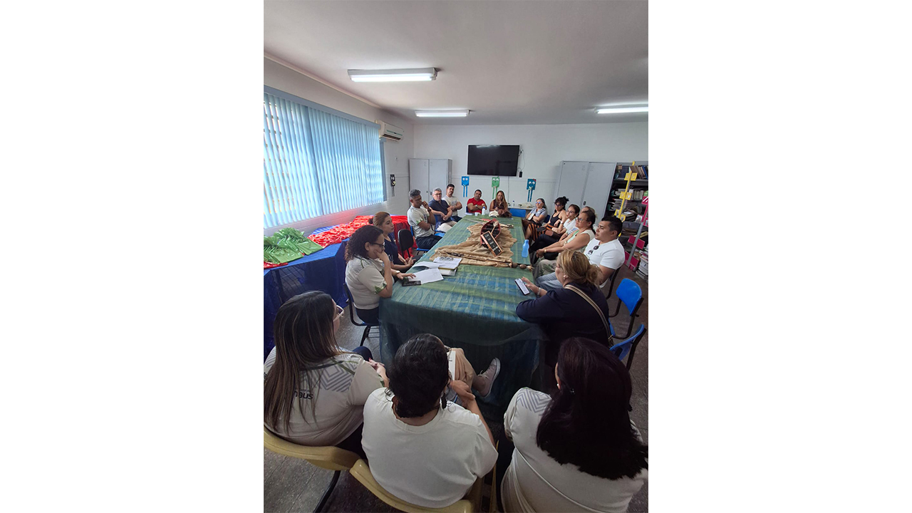 Cerca de quinze adultos de diferentes grupos raciais, vestindo roupas casuais, estão sentados ao redor de uma grande mesa. A mesa central está coberta por um tecido verde listrado e exibe uma miniatura de canoa de madeira com remos. O ambiente possui piso cinza, janelas com persianas azuis à esquerda, paredes brancas e uma TV fixada na parede ao fundo.