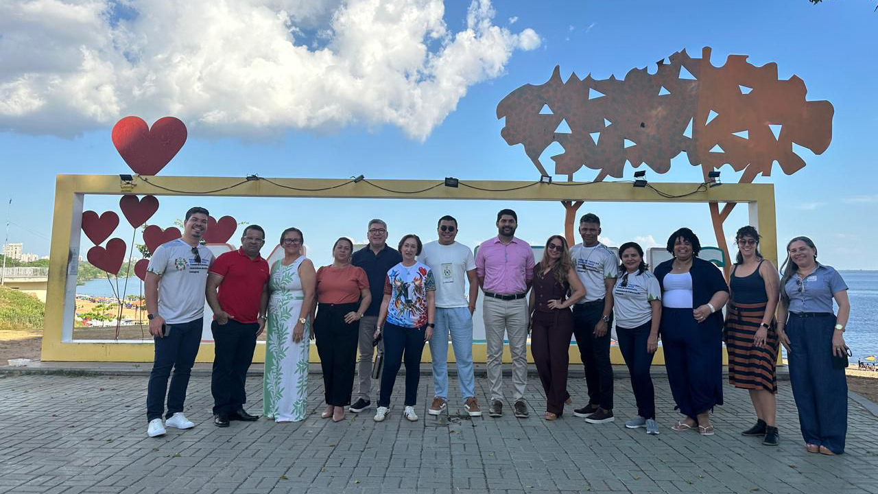 Um grupo multirracial de quatorze adultos, com roupas casuais, está em pé, lado a lado, posando para a foto. O destaque central é uma estrutura amarela atrás do grupo, decorada com corações vermelhos e uma escultura metálica em formato de árvore. Ao fundo, veem-se um rio, vegetação às margens e construções distantes.