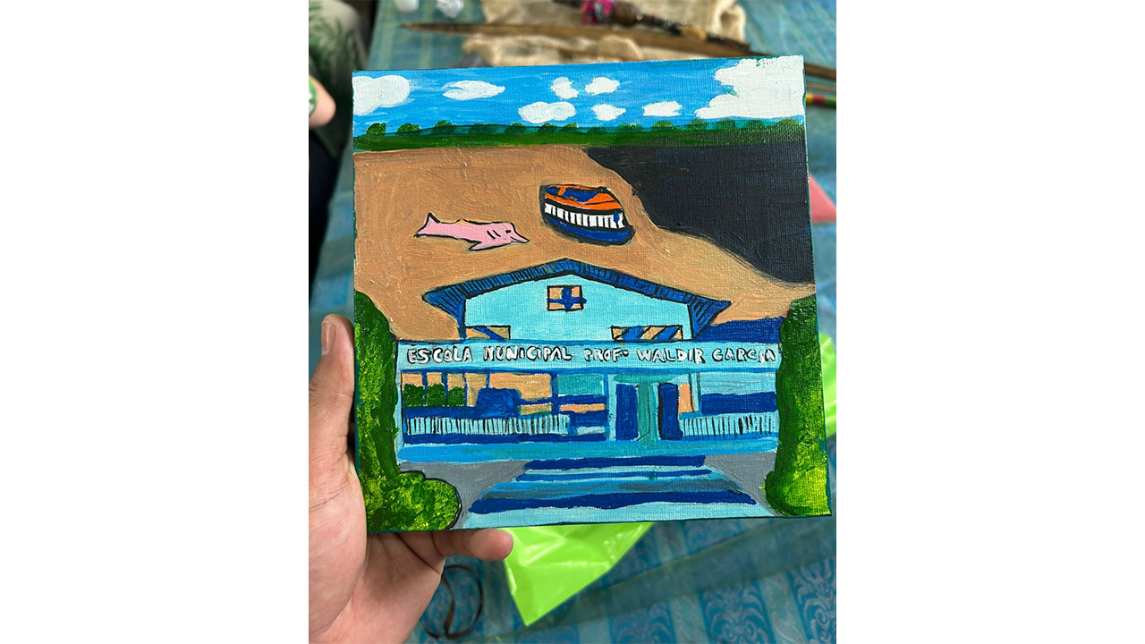 Uma pequena tela de pintura quadrada é segurada por uma mão. A pintura destaca uma fachada azul, um boto-cor-de-rosa e um barco sobre um rio. O cenário ao fundo está desfocado e mostra partes de uma mesa coberta por uma toalha plástica esverdeada.