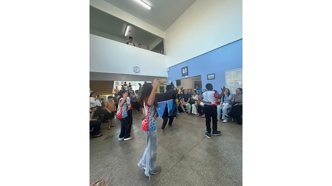 Um grupo multirracial de jovens, vestindo blusas estampadas, dança com os braços erguidos ao redor de uma figura de boi-bumbá, enquanto é observado por adultos sentados. O ambiente possui piso cinza, paredes pintadas de azul e branco, um relógio e espectadores que assistem à apresentação a partir de um andar superior protegido por tela.