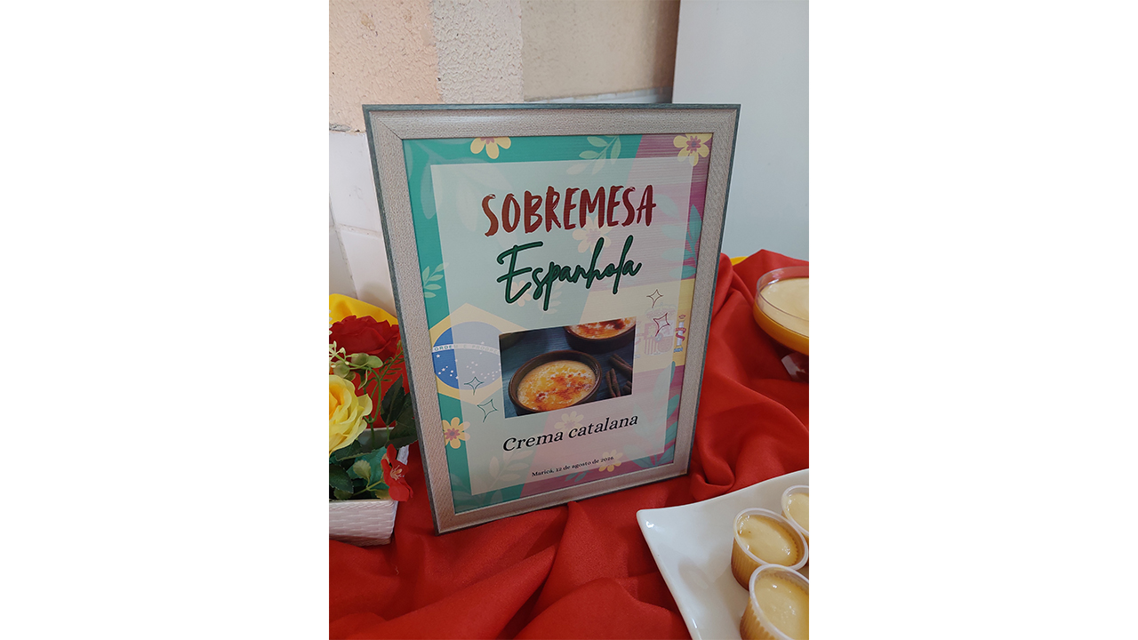 Mesa de exposição coberta com tecido vermelho. Ao centro, um porta-retrato com molduras prateadas exibe o texto “Sobremesa Espanhola – Crema catalana”, acompanhado de uma foto do doce. Ao redor, há flores decorativas e pequenos copinhos descartáveis com a sobremesa. Ao fundo, vê-se uma parede clara.