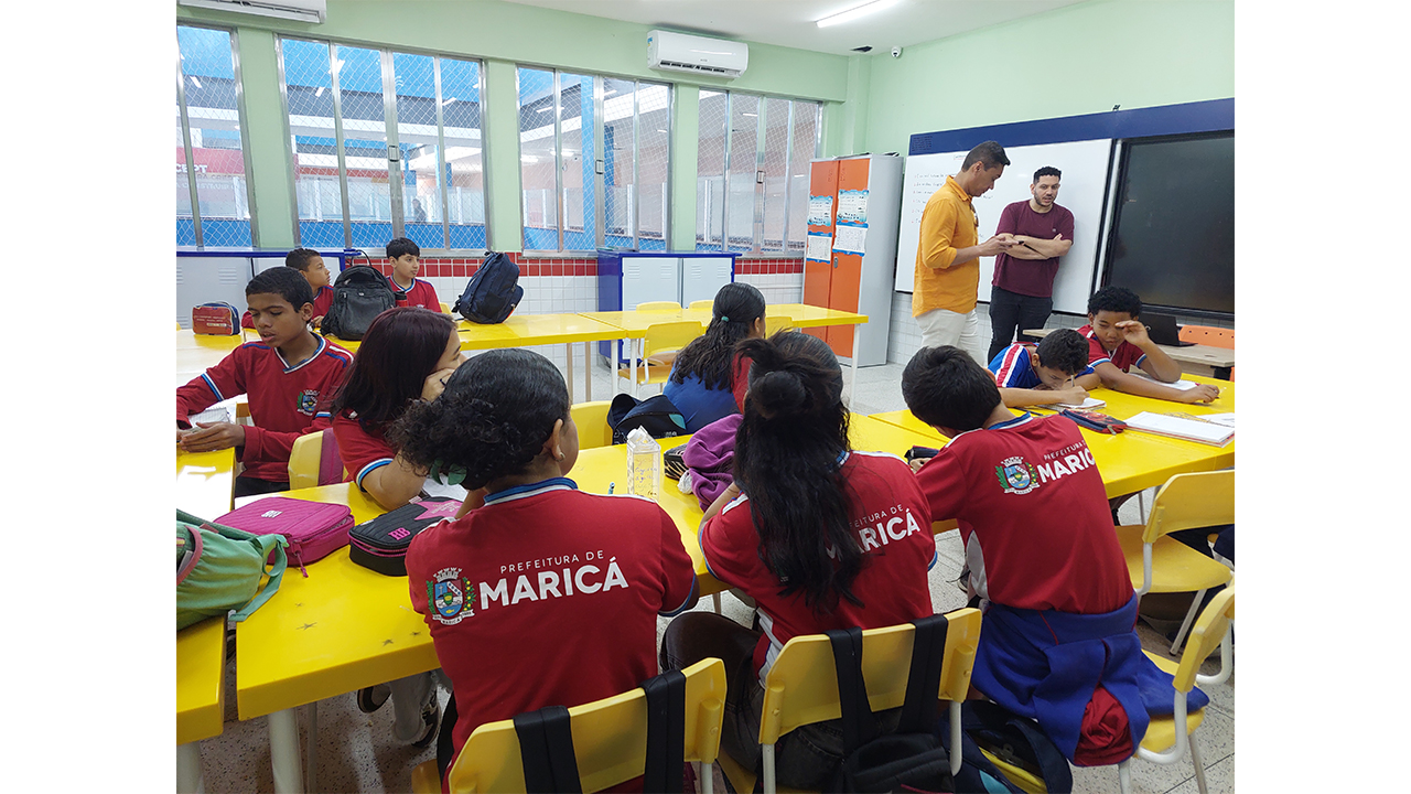 Sala de aula com estudantes sentados em mesas e cadeiras amarelas e dois adultos em pé à frente. Os alunos vestem roupas vermelhas e azuis, com a inscrição "Prefeitura de Maricá" nas costas e o brasão da cidade, enquanto os adultos conversam entre si próximos a um quadro branco e prato na frente da sala. Ao fundo, há janelas amplas e armários coloridos. As paredes da sala são da cor verde claro.