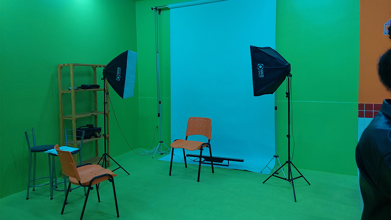 Sala verde preparada para registro fotográfico. Ao centro, há um fundo fotográfico branco, duas luzes de estúdio sobre tripés e duas cadeiras laranjas posicionadas, uma de costas para o fundo branco, centralizada, e outra de frente para a outra cadeira. À esquerda, observa-se uma estante de madeira com equipamentos.
