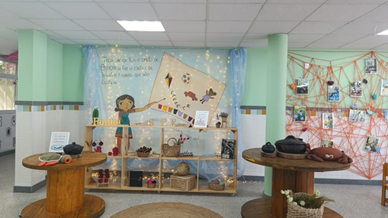 No centro de uma exposição escolar, há artesanatos sobre carretéis e uma estante de madeira, à frente de um painel ilustrado. O desenho conta com uma menina segurando um pincel, pintando uma pipa, uma bola de futebol, uma boneca e um carrinho em uma tela. À direita, fotos estão presas em uma teia de barbantes laranjas, fixada em uma parede verde-clara. O piso do ambiente é cinza.