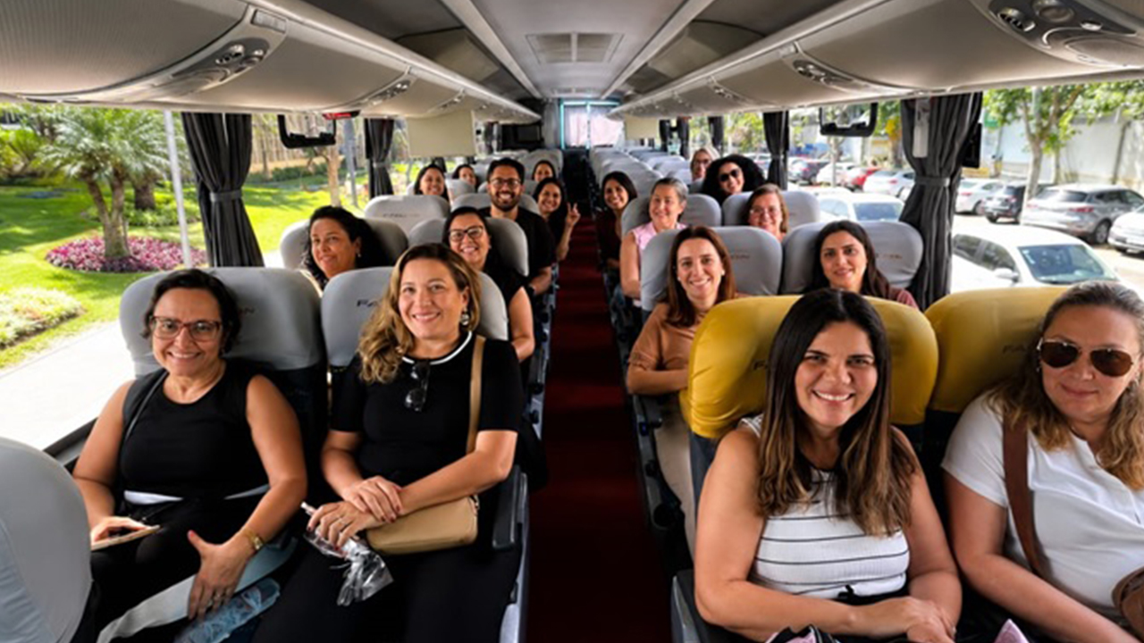 Interior de um ônibus de turismo com cerca de 18 passageiras adultas, vestindo roupas casuais, sentados e sorrindo. O corredor central divide as fileiras de poltronas. As janelas laterais revelam um cenário com gramado, árvores e carros estacionados.