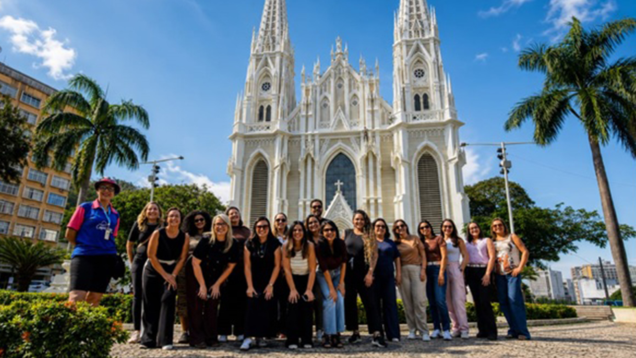 Grupo multirracial de cerca de vinte adultos posando lado a lado em uma praça. As pessoas vestem roupas casuais e sorriem. Ao fundo, destaca-se uma grande catedral, com detalhes da fachada em branco e duas torres altas, cercada por palmeiras.