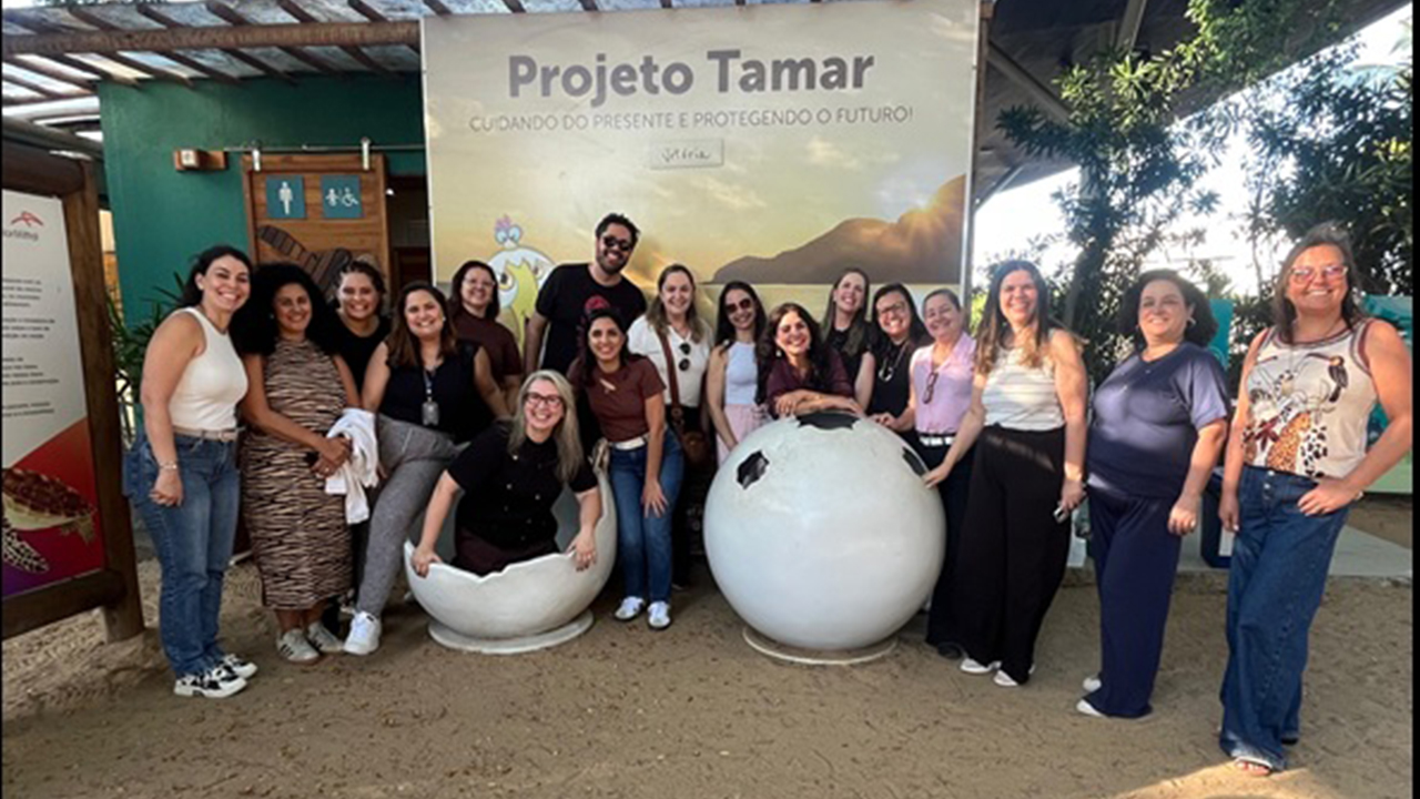 Grupo multirracial de 17 pessoas posa lado a lado, sorrindo, em volta de estruturas em formato de ovos grandes, sobre um chão de areia. Há uma mulher sentada dentro de meia casca de ovo. Atrás do grupo, destaca-se um painel do "Projeto Tamar", fixado em uma construção verde, com estruturas de madeira no telhado.