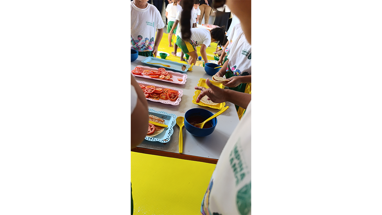 Um grupo multirracial de crianças está em pé, vestindo camisetas brancas idênticas, e interagindo ao redor de uma mesa. Elas observam e manuseiam bandejas de plástico coloridas, onde há rodelas de tomate e tigelas com molho. Ao fundo, um piso de EVA amarelo e outras crianças.