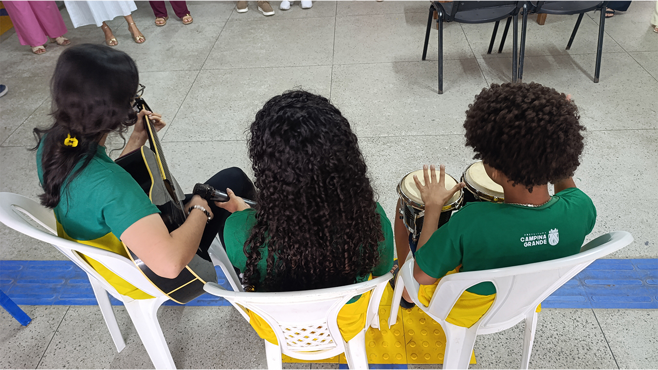 Um grupo multirracial de três jovens vistos de costas faz uma apresentação musical. As(os) estudantes estão sentadas em cadeiras plásticas brancas, de costas para a câmera, vestindo camisetas verdes e amarelas idênticas, com a inscrição "Campina Grande" em branco. Da esquerda para a direita, elas tocam violão, seguram um microfone e tocam pequenos tambores (bongôs). O piso é de cerâmica clara, com faixas de piso tátil azul.