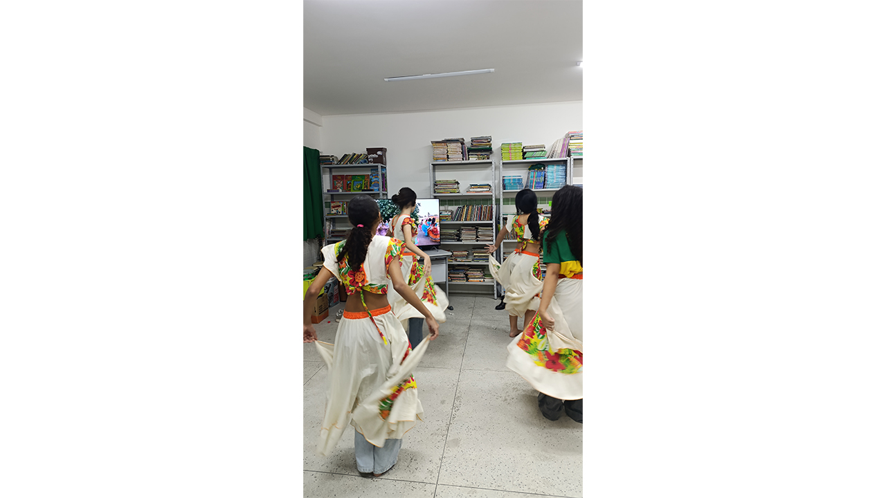 Grupo multirracial quatro jovens, vistos de costas, dança segurando as bordas de longas saias brancas, combinadas com blusas curtas com estampas florais. À frente, uma televisão exibe um vídeo de dança semelhante. O cenário inclui piso claro, cortinas verdes e uma estante de metal cheia de livros à direita.
